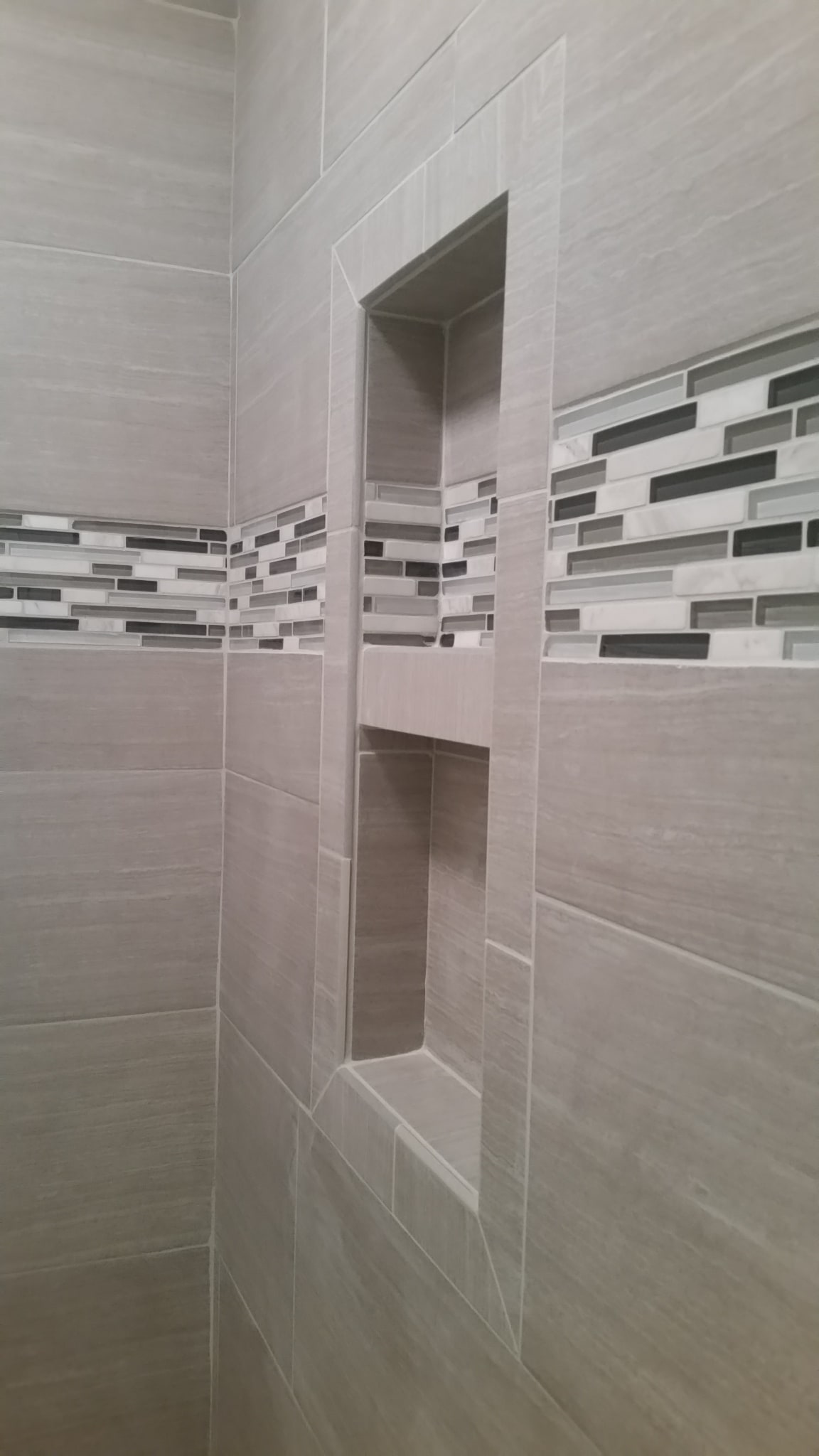 Tile shower with grey linen-look walls, vertical glass mosaic accent strip, and recessed niche shelves