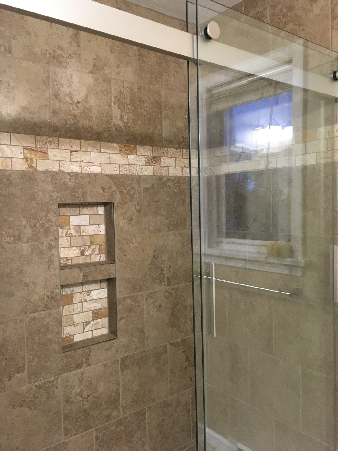 Tile shower with travertine walls, glass enclosure, natural stone mosaic accent band, and double niche