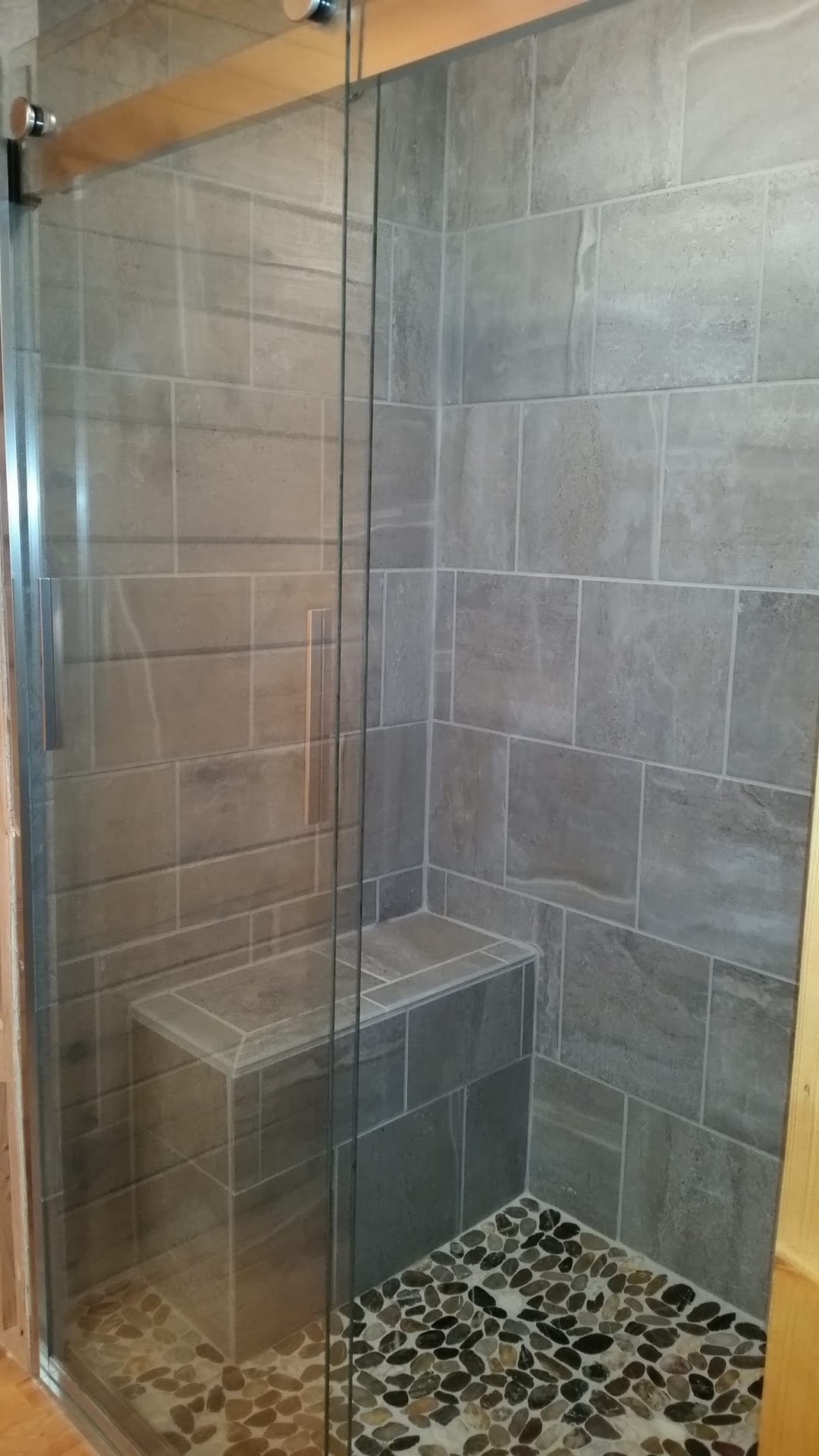 Tile shower with grey slate-look walls, built-in tile bench, pebble mosaic accent strip, and glass enclosure
