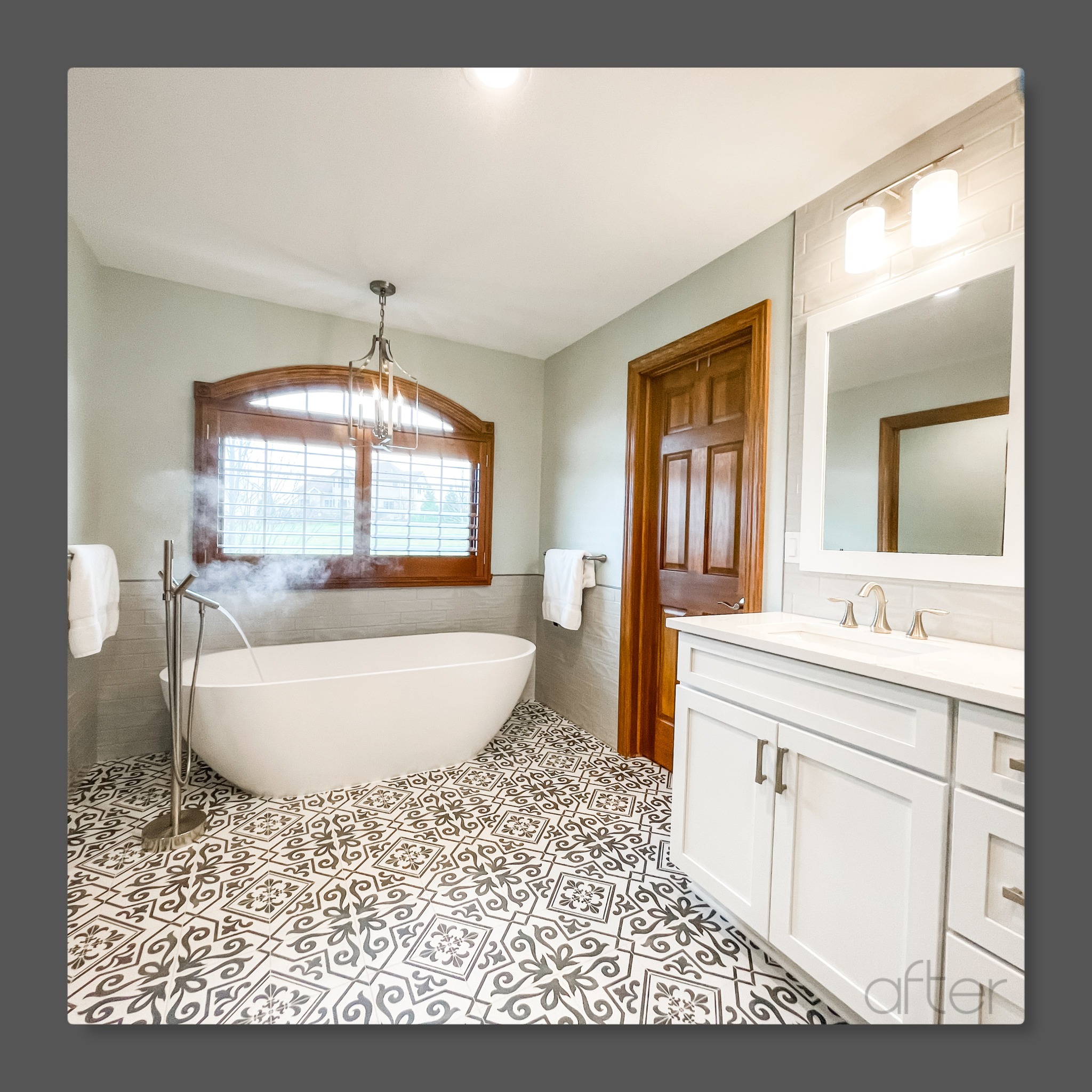 Freestanding soaking tub with floor-mounted filler, sage-green walls, patterned cement tile floor, white vanity, and lantern chandelier, Edinboro