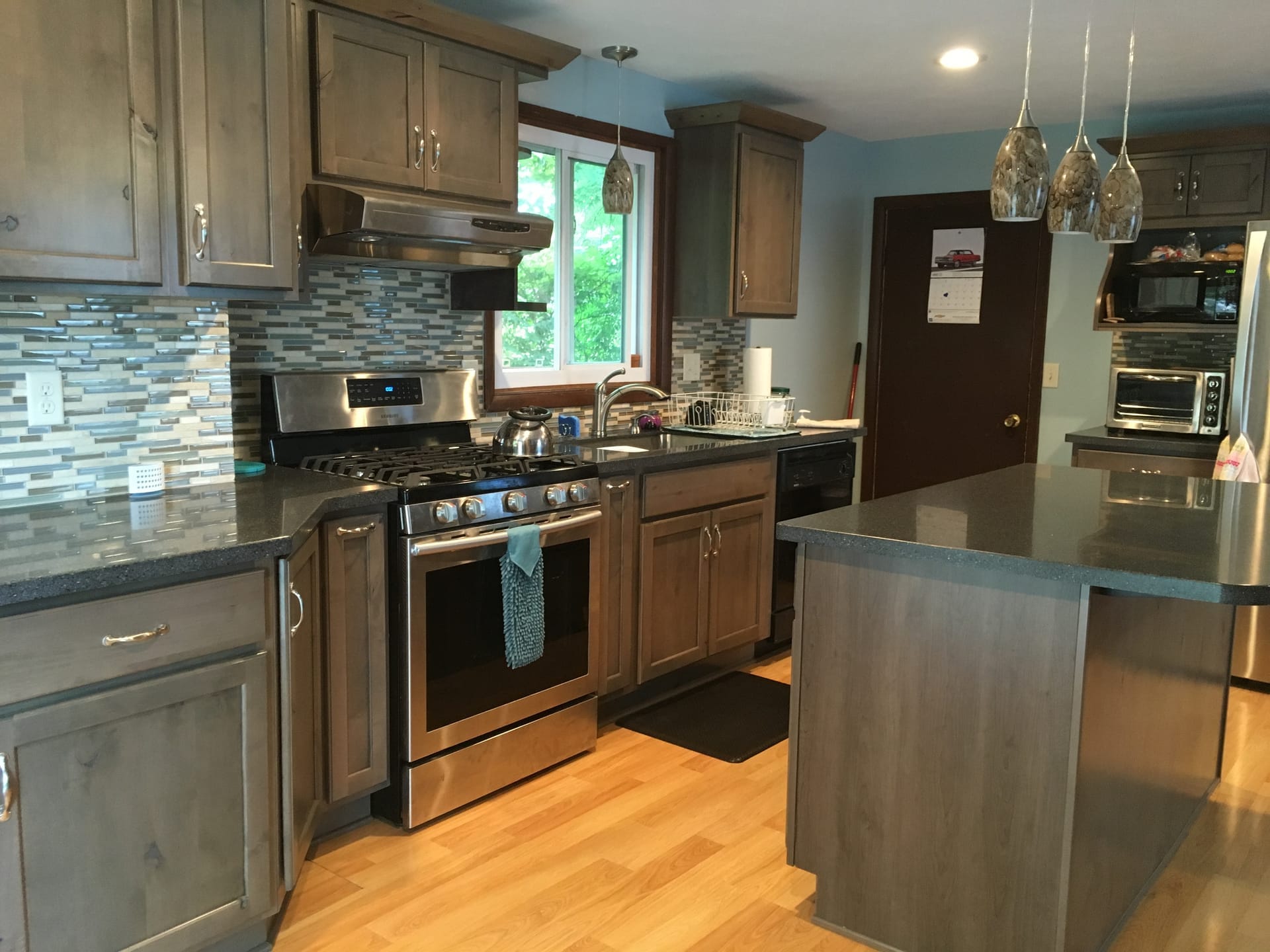 Grey-stained wood kitchen with linear glass mosaic backsplash, dark counters, center island, and light hardwood flooring, Edinboro