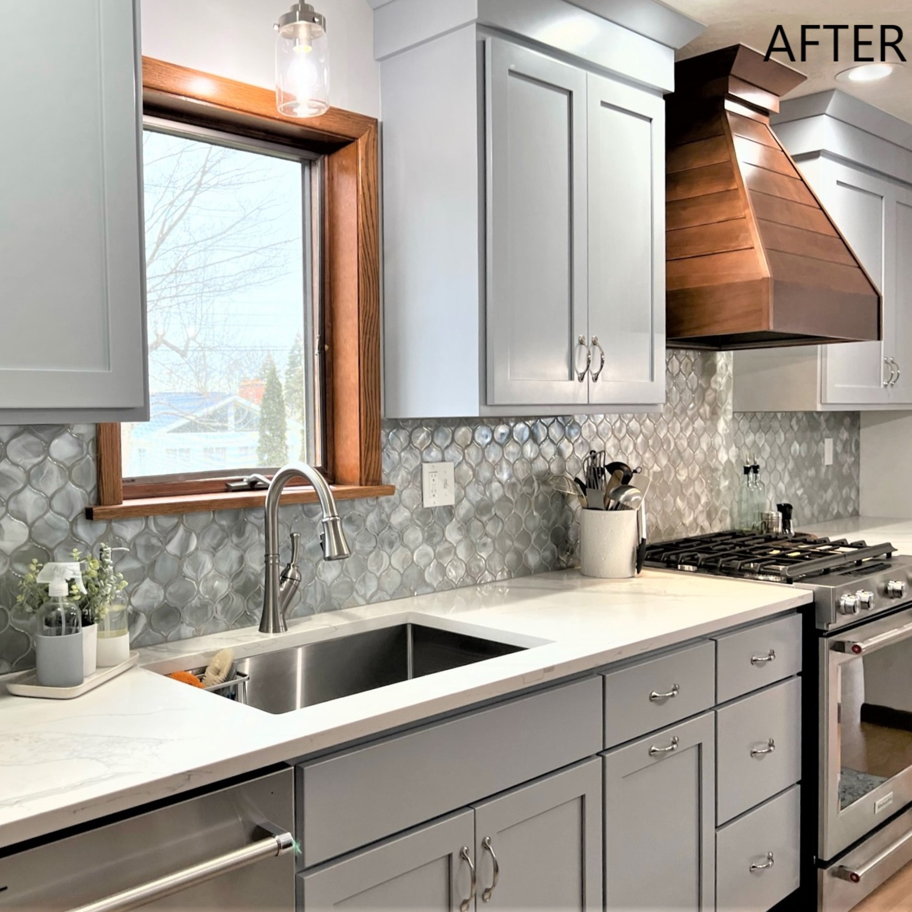 Light blue-grey shaker kitchen with white quartz counters, iridescent lantern glass tile backsplash, and dark wood range hood