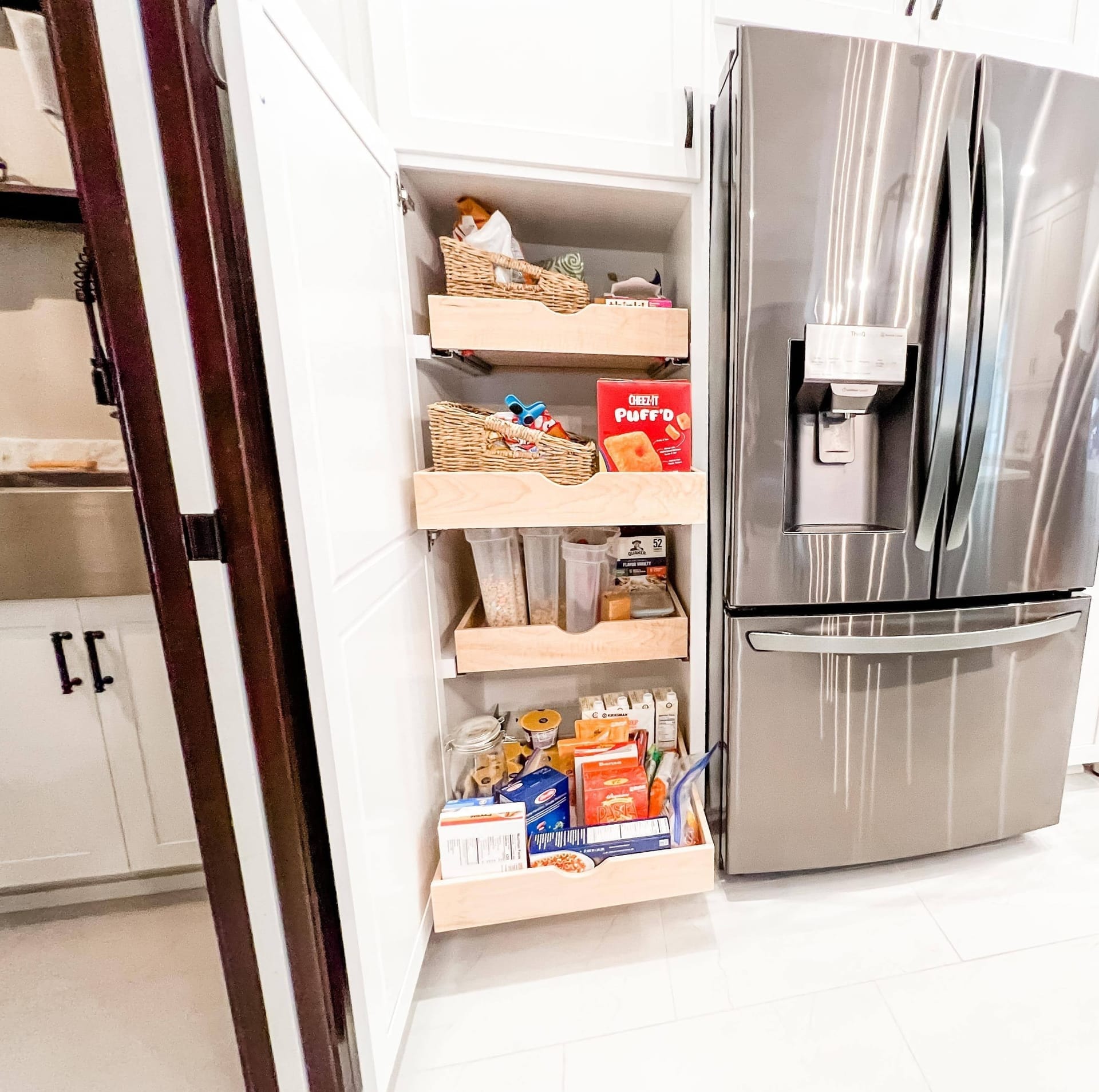 White pantry cabinet with roll-out shelving beside a stainless french-door refrigerator, tile flooring