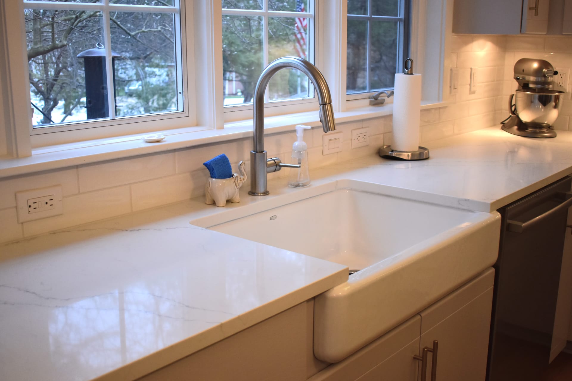 Farmhouse apron sink with marble-look quartz counters, white subway tile, and gooseneck faucet beneath triple windows