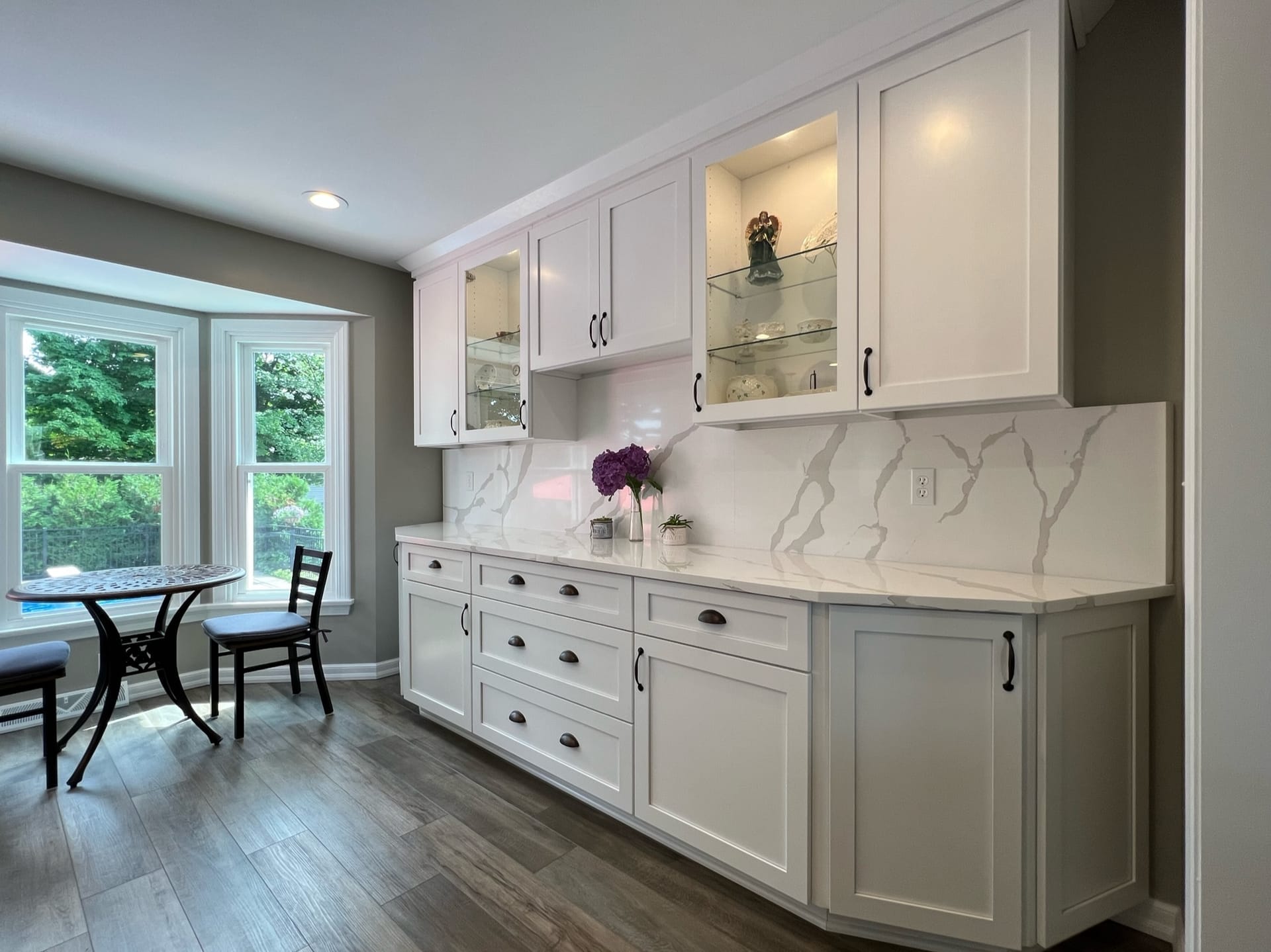 White shaker kitchen with glass-front cabinets, marble-look quartz counters, slab backsplash, and bay-window dining nook