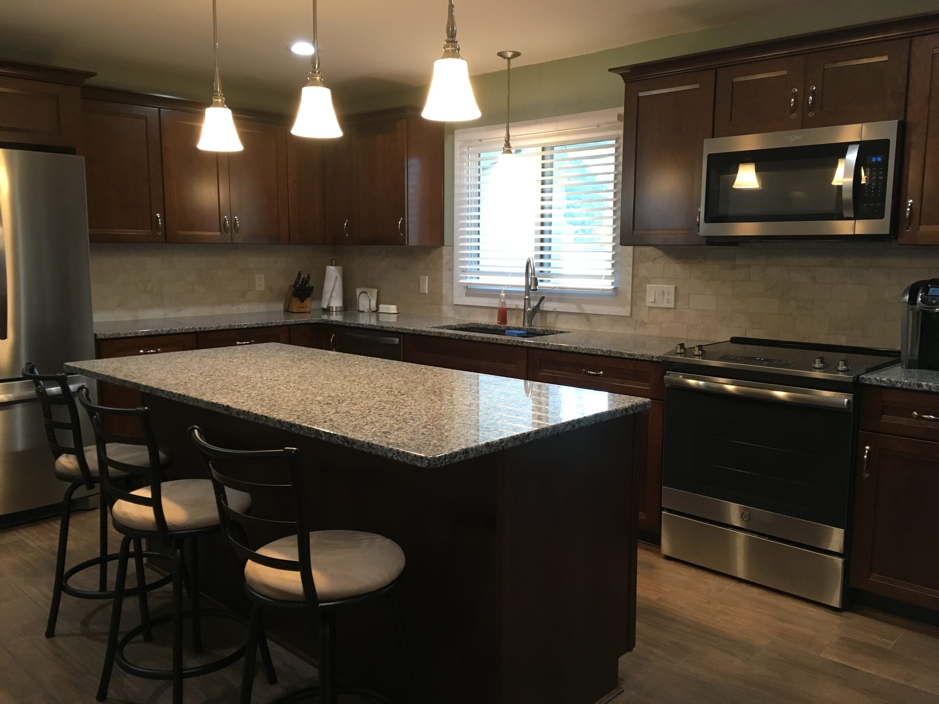 Espresso shaker kitchen with travertine subway tile, speckled granite island, stainless appliances, and glass pendant lighting