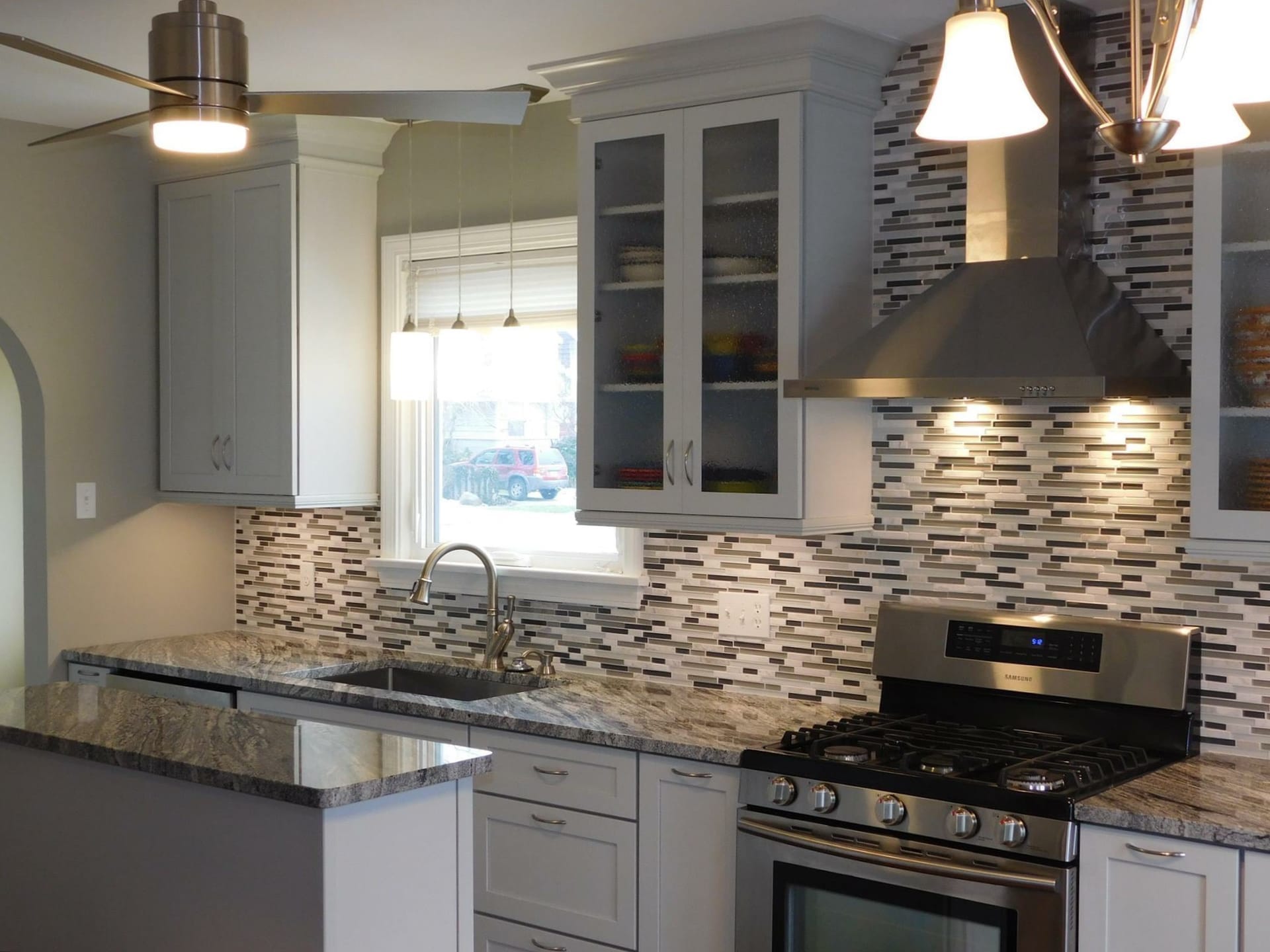 Grey shaker kitchen with linear stone mosaic backsplash, speckled granite counters, stainless chimney hood, and glass-front cabinets
