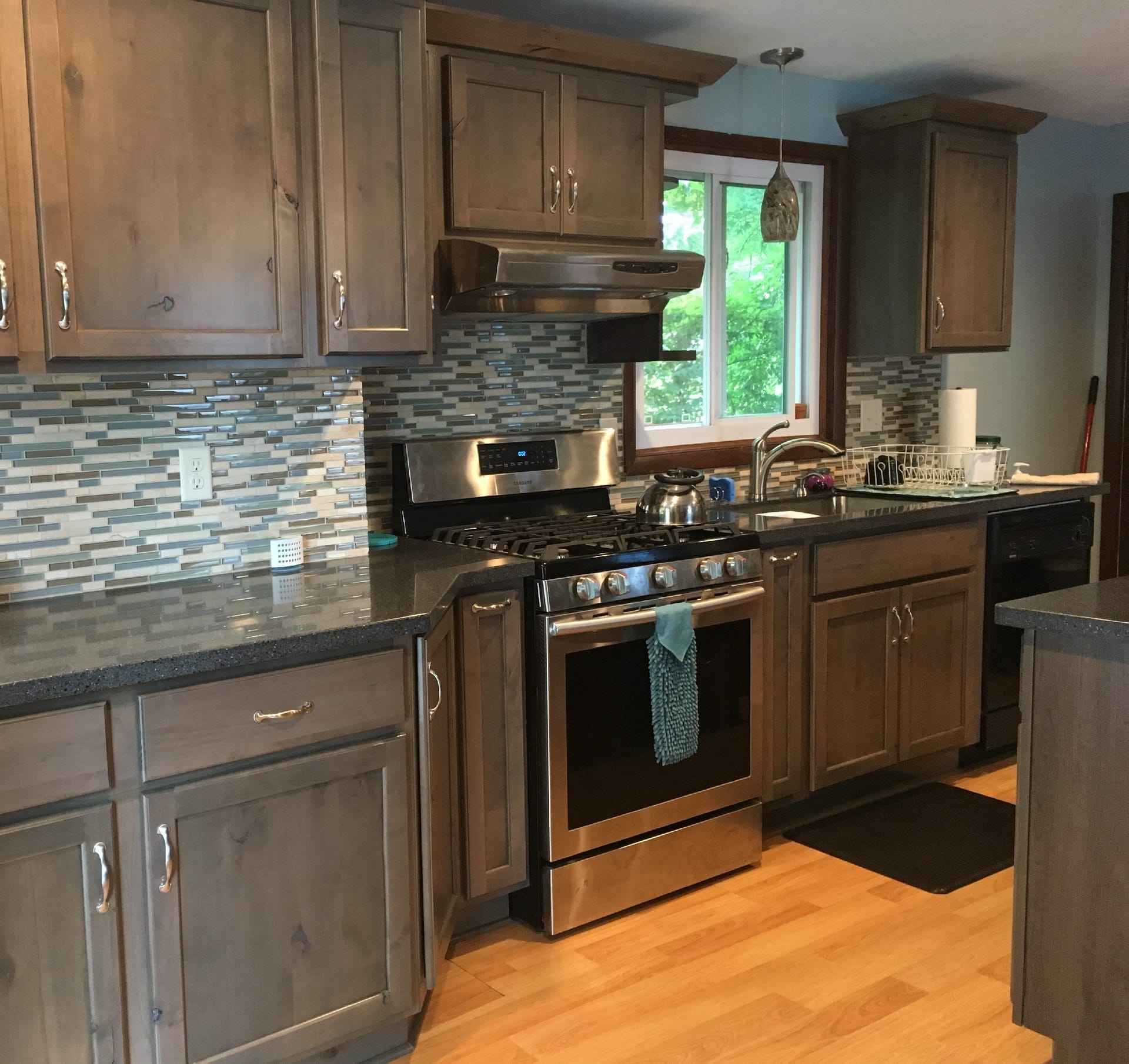 Grey-stained wood kitchen with linear glass mosaic backsplash, dark granite counters, stainless gas range, hardwood flooring, Erie PA