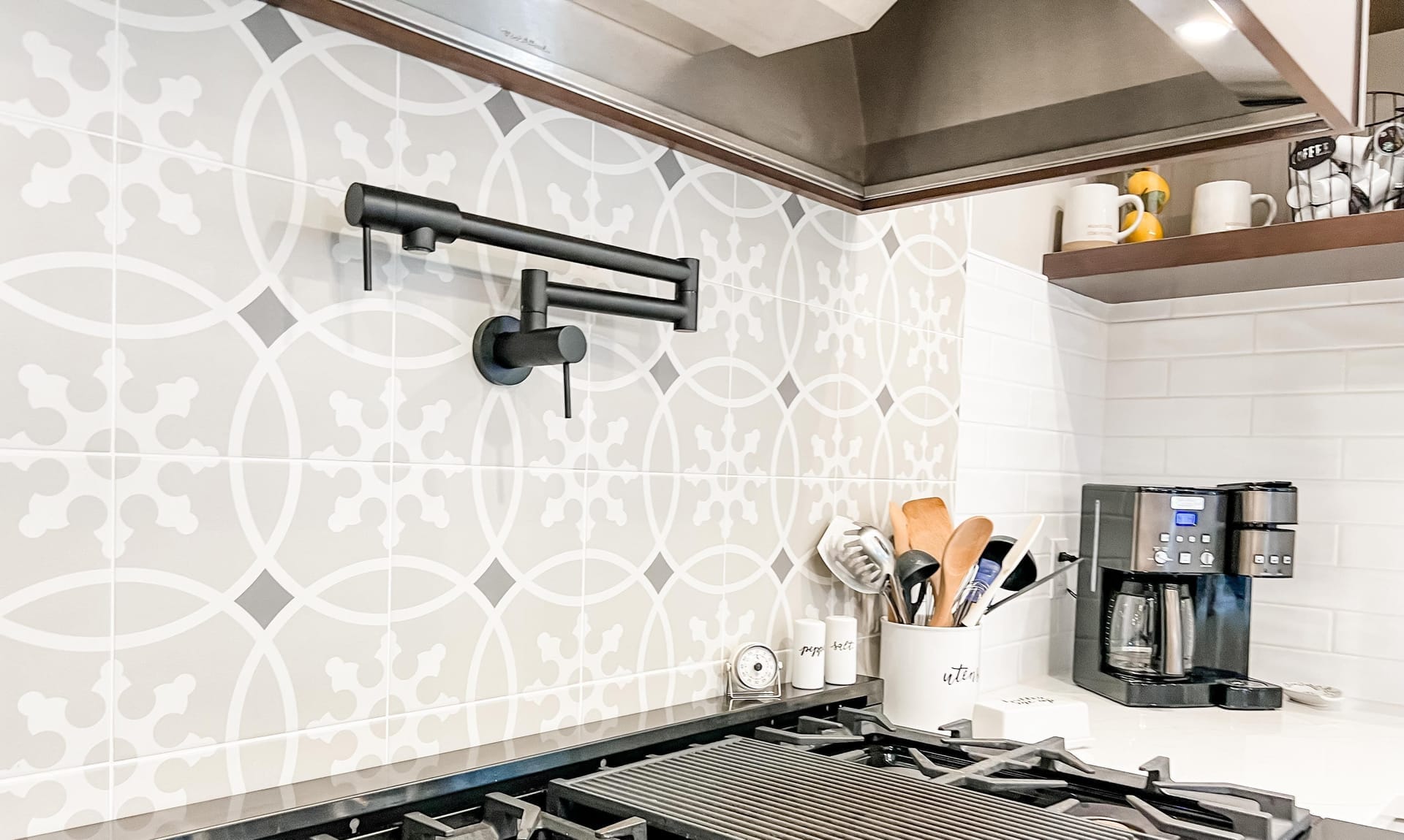 Grey patterned cement tile backsplash with matte-black pot filler, wood range hood, and professional gas range
