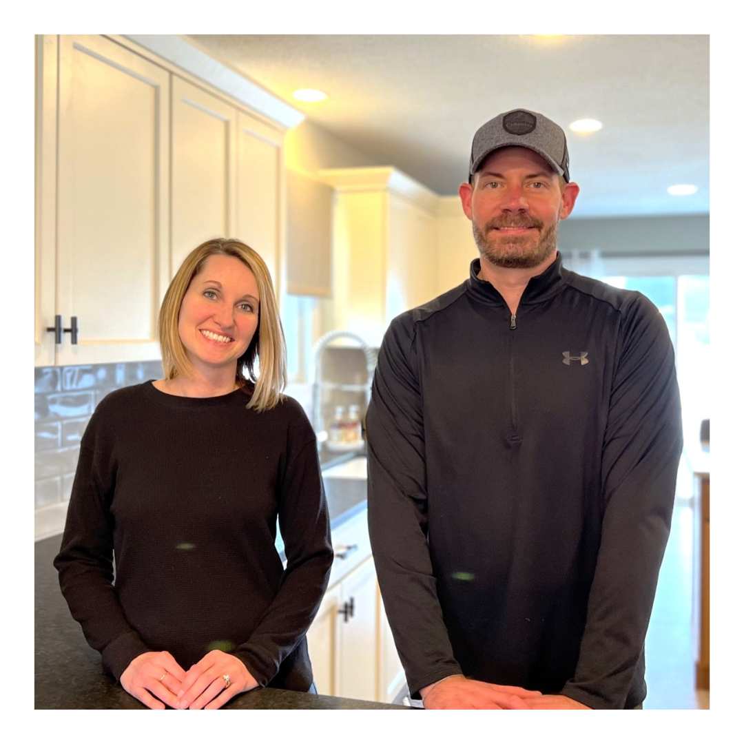 Two people standing in a white cabinet kitchen with grey subway tile backsplash and dark counters
